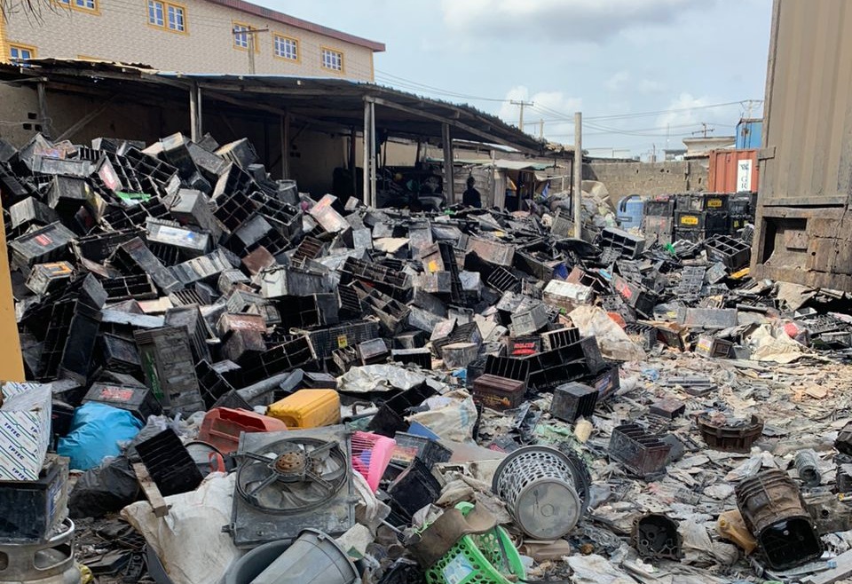 A used lead-acid battery draining and breaking site in Lagos, Nigeria, with a large heap of discarded black plastic battery casings piled across the yard.