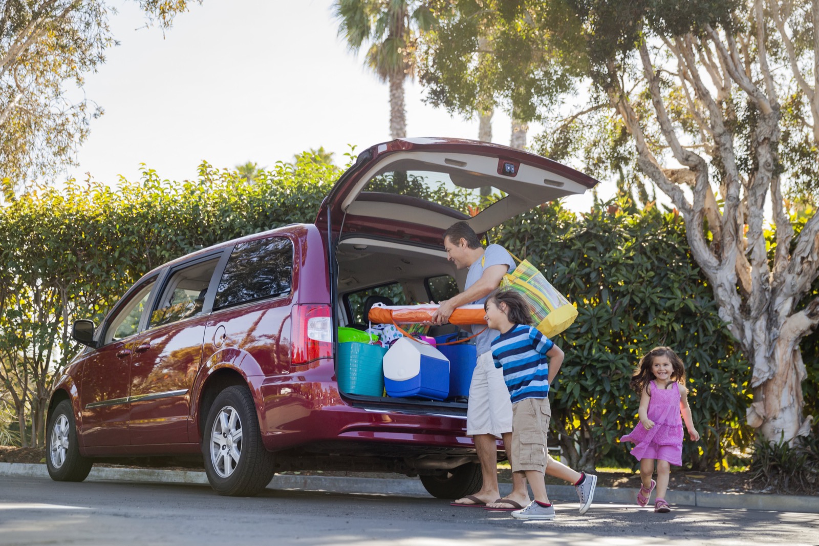 A father loads coolers and beach gear into the open rear hatch of a red minivan parked in a driveway as his two young children walk up to join him.